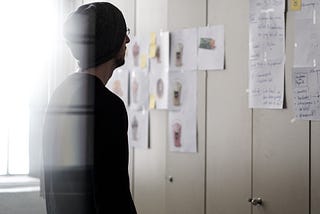 Man in beanie and glasses faces away from the camera looking at a wall with pieces of paper taped to it. The image is over exposed on the left edge with sunlight coming through the window
