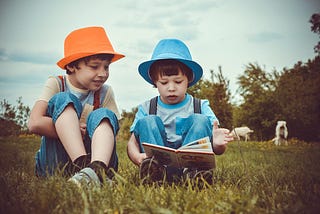 Two boys sitting on grass. One holds a book and the other one is looking at the book and smiling.