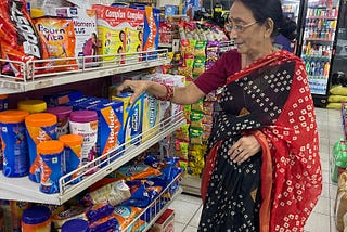 A woman in a tie-and-dye flowing garment is shopping in the Horlicks (processed malt-based powder) aisle at a grocery store. The woman is wearing bangles. There is a strip of masala packages hanging behind her.