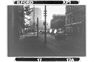 A 1980s urban street, gritty in black and white, cars sit at the light, the sun silhouettes the buildings and passers by