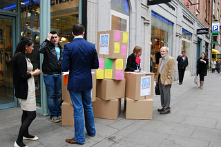 Two researchers standing behind a stack of cardboard boxed with posters reading ‘help us design a better grafton quarter’ in Dublin. Each researcher is talking to a member of the public.