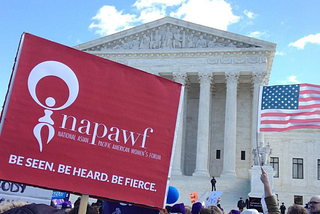 Red NAPAWF sign saying “Be Seen. Be Heard. Be Fierce.” lifted above crowd in front of the Supreme Court