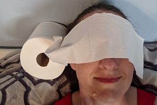 Close-up of a white woman lying against a purple blanket with a roll of toilet paper next to her head, the loose end draped across her eyes