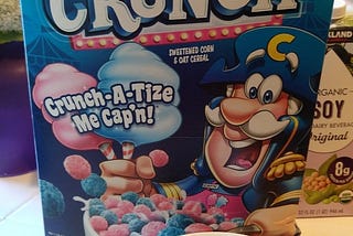 a box of “Captain Crunch Cotton Candy Crunch cereal”, with a bowl of the cereal in front of it. Both are blue and pink.
