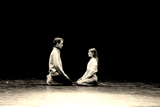 Two actors kneel facing each other on an empty, minimalist stage. They appear calm, focused and open to each other, ready to listen thoughtfully.