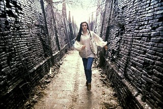 A scene from the movie Labyrinth: A young, white woman in a white poet’s blouse, vest, and jeans runs along a desolate labyrinth hallway with broken bricks and dead vines on an overcast day.