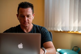 An adult man with a pensive expression and spiky black hair is sitting at his Macbook laptop, wearing white earbuds.