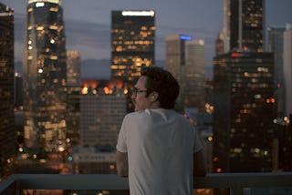 Man in white shirt overlooks city buildings and lights poignantly.