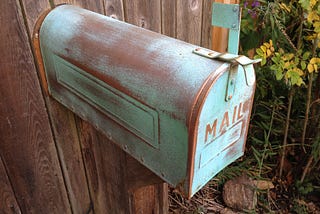 Photo of an old, worn mailbox, green patina or paint, with its flag up, attached to a wooden fence. The word MAIL is on the front flap.