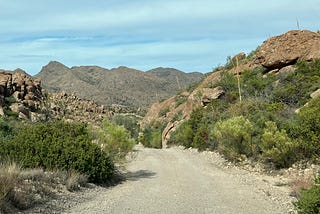 A dirt road in the desert winds between rock formations with mountains in the background.