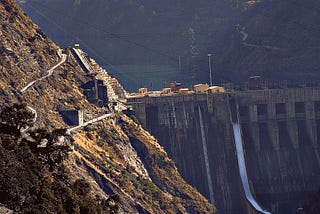 A steep-sided valley with small trees on the hillsides is shown, with sunshine on one slope, and Baglihar Dam mostly in shadow. A white stream of water flows through one of the dam’s sluice gates, and a small amount of the dam’s reservoir is visible in the background. Baglihar is a run-of-river power project on the Chenab River in the southern Doda district of the Indian state of Jammu and Kashmir. This is a closer crop of the photo included in the body text of the article.