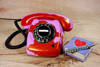 A red and pink rotary phone with a small notepad and bejeweled pen beside it.