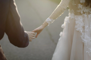 A couple loosely holding hands, one wearing a white gown with floral details, the other in a dark suit, under soft lighting.