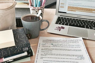 a partially visible laptop kept on a table alongside papers, notebooks, a cup of pens, and a cold espresso.