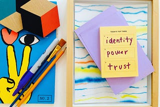 Tableau of a colorful magazine cover featuring an illustration of a hand making a peace sign, an eye, and a heart. A pencil, Sharpie, and paint brush lay on top of the magazine. A post-it with a hand-written list “ identity, power, trust” rest atop a framed piece of colorful line art.