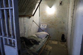 Small, dirty, prison cell with a bed and urinal. It looks very depressing.