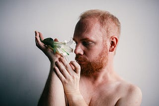 A white topless man with a beard holding a white flower in his hand.