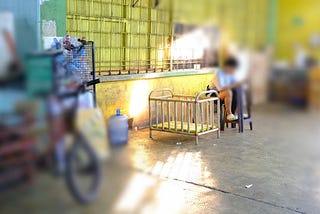 A rattan baby crib sits in a well-lit corner of an open market in Cebu City, Philippines. Sunlight casts a warm glow on the crib, highlighting its simplicity. Nearby, a person sits on a chair, partially visible, while the surrounding area reflects the bustling yet rustic ambiance of the market with scattered items and vibrant colors.