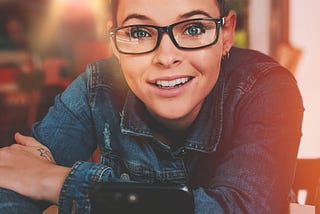 Woman wearing Demin, sitting in the public place like cafe with a phone in her hand, and a stylish hairstyle. She is looking quite happy and attentive. Maybe thinking about the 11 Marketing AI tools to stay ahead of 99% of people! Photo by Alex Perez on Unsplash.