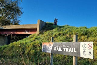 A sign saying ‘Gippsland Plains Rail Trail’ in front of a grassy hill and old rail overpass