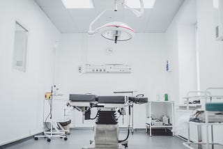 A photo of a surgical room. A dark bed is in the middle, on a hydraulic lift. A large lamp is positioned over the bed. A variety of tables and carts with supplies sit around the bed. The walls are white and the floor is gray.
