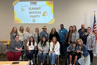 A group of 20 people stands or sits in front of a projector screen that has the words “Summit Watch Party” displayed.