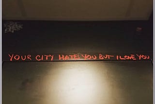 red graffiti on a black wall in a bathroom that says, “your city hates you but I love you.”