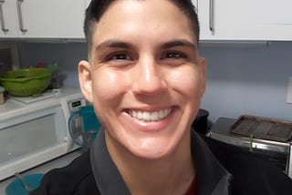 A selfie of the writer, Rox Trujillo. She is smiling, teeth showing, has short dark brown hair, eyebrows, and eyes. Her hair cut is medium side fade, long on top, combed towards one side.