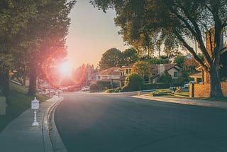 A view looking down a tree-lined road in a suburban neighborhood with houses on either side and the sun just above the horizon