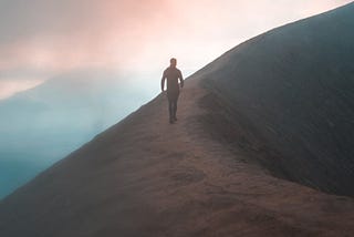 A person walking alone on a narrow, winding mountain ridge surrounded by mist and soft pink-blue dawn light, evoking exploration and solitude.