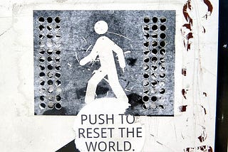 A sticker on a crosswalk signal button that reads “Push to reset the world” in all caps