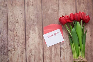 Against a background of wooden boards, a twine-wrapped bundle of red tulips sits beside a small hand-written note that reads “Thank you,” paired with a red envelope.