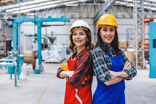 Two young women standing back to back, wearing hard hats and standing in an engineering facility.