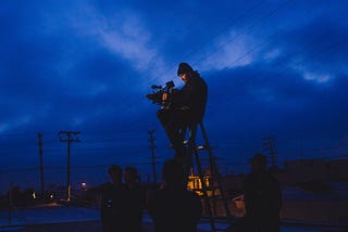 A camera operator sits on a stepladder against a blue night sky