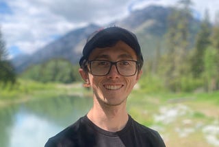 A man in his mid 20s with a black cap and glasses smiling. He has a black t-shirt. In the background, a river is flowing with mountains rising above the horizon.