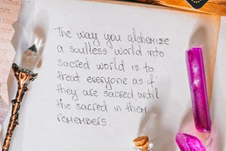 Several crystals and a sheet of paper with the writing: “The way you alchemize a soulless world into a sacred world is to treat everyone as if they are sacred until the sacred in them remembers.”