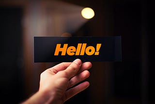 One hand shows a sign which has a black background and orange writing. On the sign is written “Hello”.