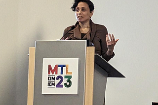 Kelly Bron Johnson stands at a podium delivering a talk. They are a light-skinned Black person with a crop of short, wavy, dark hair combed to one side. They are mid speech and talking with their hands, one of which is raised, palm up.