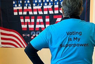 My colleague Lois facing a flag that says “Save Democracy Harris Walz” wearing a blue t-shirt that says “Voting is My Superpower.”