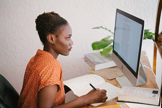 A black woman in an orange blouse with white dots staring into an apple monitor over notes in a notebook.