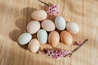 Ten chicken eggs in natural colors on a light-colored wooden surface, with two sprigs of fresh cherry blossoms at the edges of the image