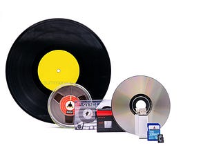 a photo of multiple items containing music, including a vinyl album, a cassette tape, a reel-to-reel tape, and a compact disc