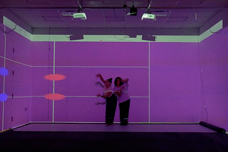 To dancers perform in a room with purple lighting, responding to projected lines and shapes on the walls. The digital visuals from a grid-like score that guides their improvised movements.
