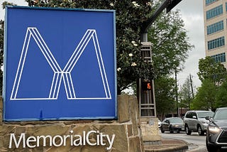 A blue sign with the capital M at a busy city intersection and a large magnolia tree in the background. At the bottom of the sign, it reads, “Memorial City.” On the right in the background is a hospital.