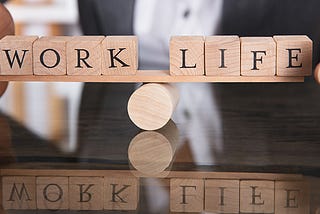 Image shows letter blocks with 4 letters on each end of a horizontal flat platform balancing on a a round cylender. The first 4 letters on the left spell ‘Work” the four letters on the right read “Life”. The image shows perfect balance.