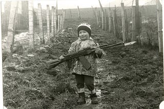 A black and white photo of a young boy of about four, the author, who stands between two barbwire fences at an Eastern European border. The boy is on the muddy pass and holding a large AK 47 gun with an afixed bayonet.