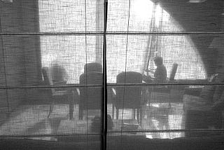 A woman sits at a window, alone, looking outside through a net curtain