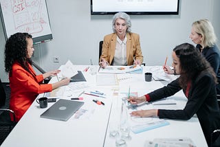 Four women of different ages sit around a conference table with documents on it and whites boards with graphs and other data written on them behind it.
