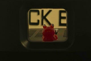 A small red toy frog with large round eyes sits centered inside a narrow rectangular opening, with blurred black letters in the background. The framing creates a tunnel-like view that draws attention to the bright, playful figure.