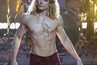 A scene from the TV series “The Vampire Lestat.” It shows actor Sam Reid as Lestat, standing on a concert stage during a performance. He has on no shirt and is wearing tight pants. A drum set is behind him.
